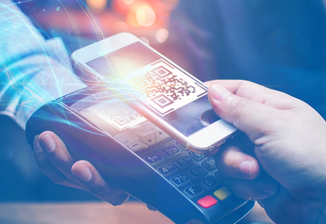 Primer plano de un pago sin contacto. Una mano sostiene un smartphone cerca de un terminal de pago que muestra un código QR.